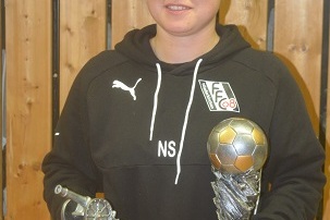 Sportlerin h&auml;lt zwei Pokale: einen mit einer Kanone und einen mit einem Fu&szlig;ball, vor einer Holzpaneelwand.