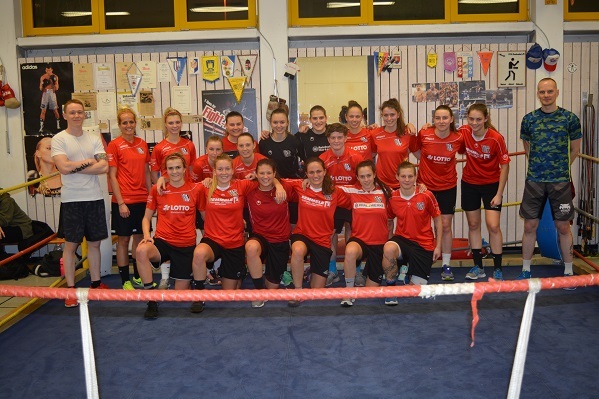 Gruppe von 15 Frauen in roten Trikots und Sportkleidung, posierend in einem Trainingsraum mit Boxring.