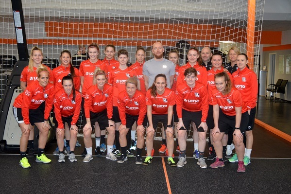 Gruppe von 20 Fu&szlig;ballspielerinnen in roten Trikots posiert vor einem Fu&szlig;balltor in einem Fitnessraum.