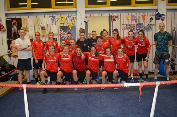 Gruppe von 15 Frauen in roten Trikots und Sportkleidung, posierend in einem Trainingsraum mit Boxring.