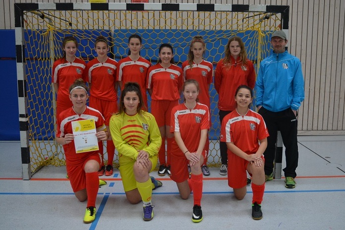 Mannschaft von jungen Fu&szlig;ballspielerinnen in roten Trikots steht vor dem Tor in einer Sporthalle, Trainer daneben.