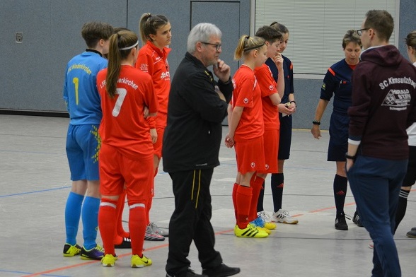 Zwei Fu&szlig;ballmannschaften in roten Trikots und eine in blauen Trikots stehen sich in einer Halle gegen&uuml;ber.