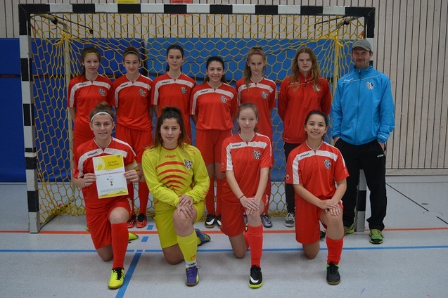 Mannschaft von jungen Fu&szlig;ballspielerinnen in roten Trikots steht vor dem Tor in einer Sporthalle, Trainer daneben.