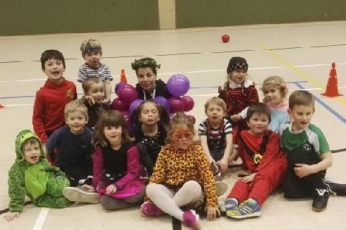 Gruppenszene von Kindern in bunten Kost&uuml;men, sitzend auf dem Boden in einer Sporthalle, umgeben von Spielgegenst&auml;nden.