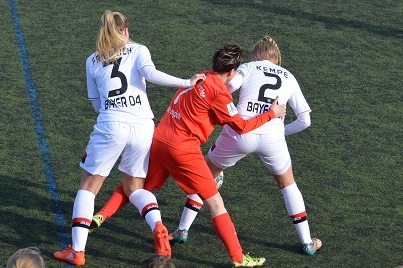 Drei Fu&szlig;ballspielerinnen in action auf dem Spielfeld, zwei in Wei&szlig; und eine in Rot, k&auml;mpfen um den Ball.