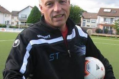 Ein Mann mit grauen Haaren h&auml;lt zwei Fu&szlig;balltore auf einem Fu&szlig;ballplatz, bekleidet mit einem schwarzen Traineranzug.