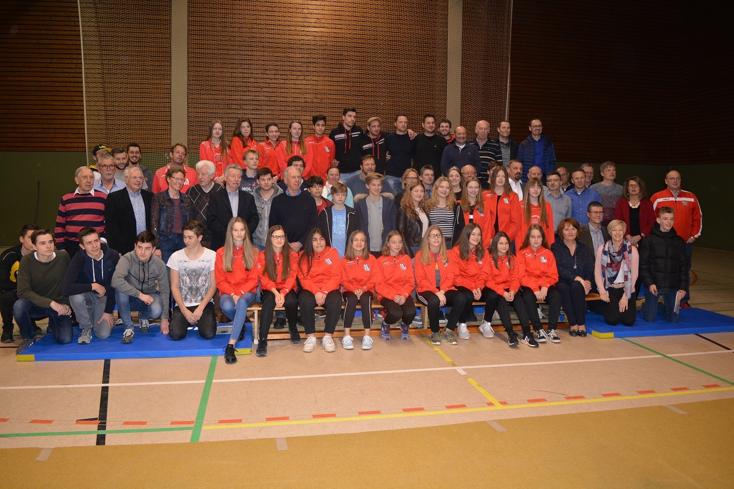 Gruppenfoto von Sportteams und Zuschauern in einer Sporthalle, alle in unterschiedlichen Outfits, auf blauem Mattenboden.