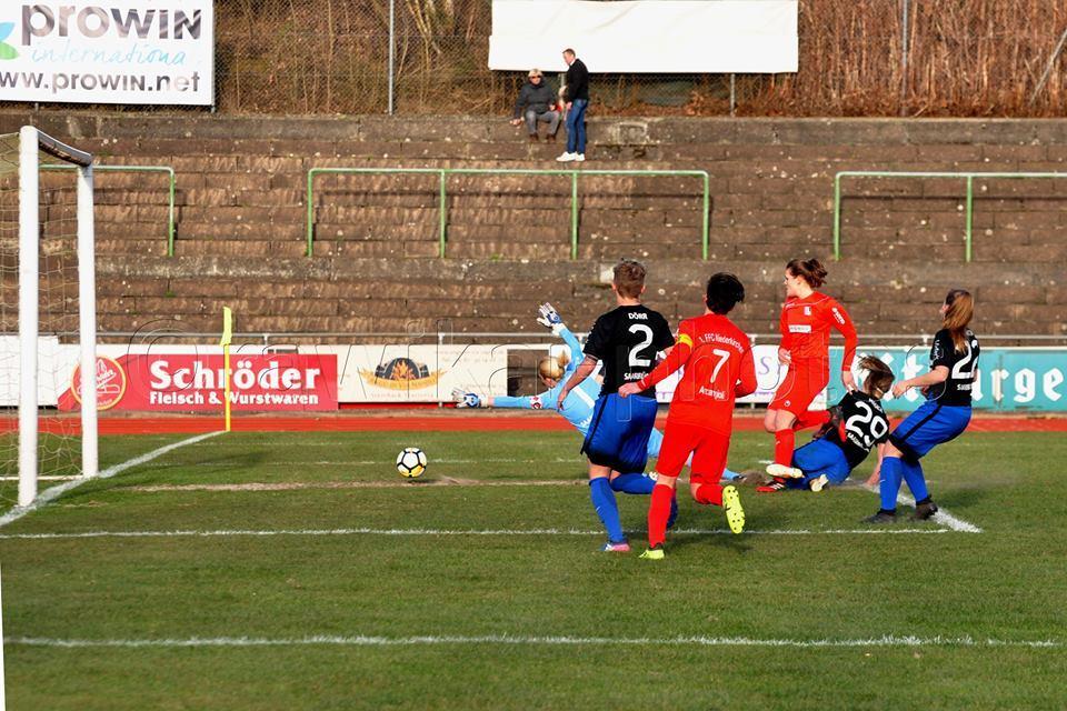 Torwart hält einen Schuss in einem Fußballspiel, während Spielerinnen beider Teams um den Ball kämpfen.