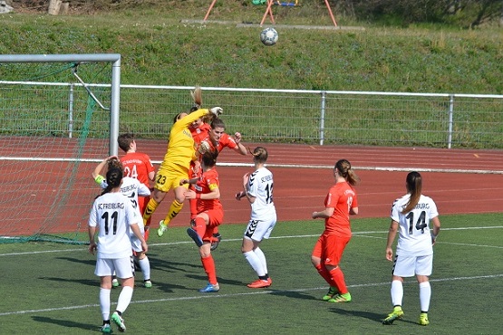Torh&uuml;terin springt in gelber Uniform, um einen Ball w&auml;hrend eines Fu&szlig;ballspiels abzuwehren, umgeben von Spielern.