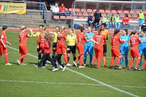 Frauenfu&szlig;ballmannschaften sch&uuml;tteln sich vor Spielbeginn auf dem Feld die H&auml;nde, Zuschauer im Hintergrund.