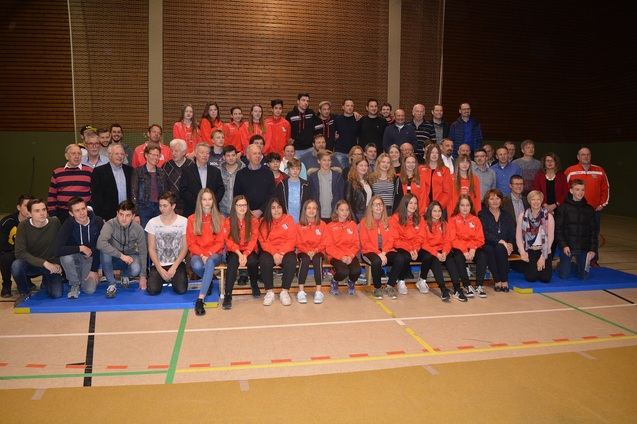 Gruppenfoto von Sportteams und Zuschauern in einer Sporthalle, alle in unterschiedlichen Outfits, auf blauem Mattenboden.