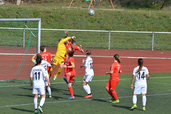 Torh&uuml;terin springt in gelber Uniform, um einen Ball w&auml;hrend eines Fu&szlig;ballspiels abzuwehren, umgeben von Spielern.