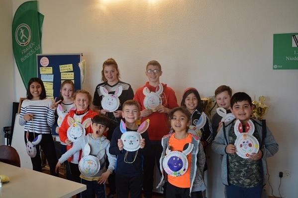 Gruppe von Kindern h&auml;lt selbstgebastelte bunte Hasenmasken in einem Raum mit Ausbildungsmaterialien.