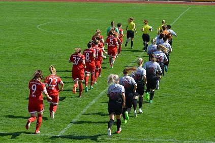 Gruppenauslauf von Frauenfu&szlig;ballmannschaften in roten und grauen Trikots auf einem gr&uuml;nen Rasenplatz.