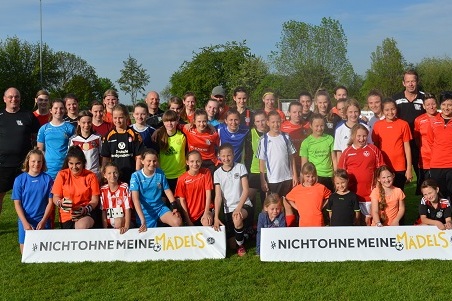 Gruppenfoto von M&auml;dchen und Frauen in Sportkleidung auf einem Fu&szlig;ballplatz, mit Banner &bdquo;Nicht ohne meine M&auml;dels&ldquo;.