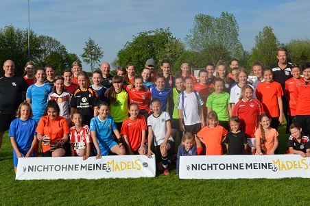 Gruppenfoto von M&auml;dchen und Frauen in Sportkleidung auf einem Fu&szlig;ballplatz, mit Banner &bdquo;Nicht ohne meine M&auml;dels&ldquo;.