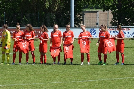 Mannschaftsaufstellung mit Spielerinnen in roten Trikots und Torh&uuml;terin, die Trikots auf dem Fu&szlig;ballplatz halten.