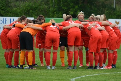 Gruppenumarmung von Spielerinnen in roten Trikots auf einem Fu&szlig;ballplatz, vorbereitet f&uuml;r das Spiel.