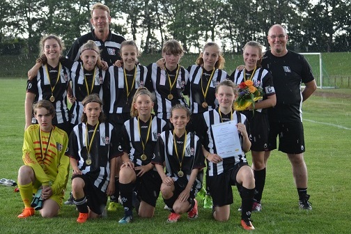 M&auml;dchenfu&szlig;ballmannschaft in schwarz-wei&szlig;en Trikots mit Medaillen, Pokal und Urkunde, posiert auf einem Fu&szlig;ballplatz.