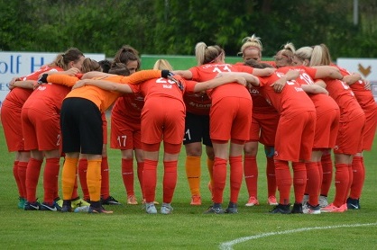 Gruppenumarmung von Spielerinnen in roten Trikots auf einem Fu&szlig;ballplatz, vorbereitet f&uuml;r das Spiel.