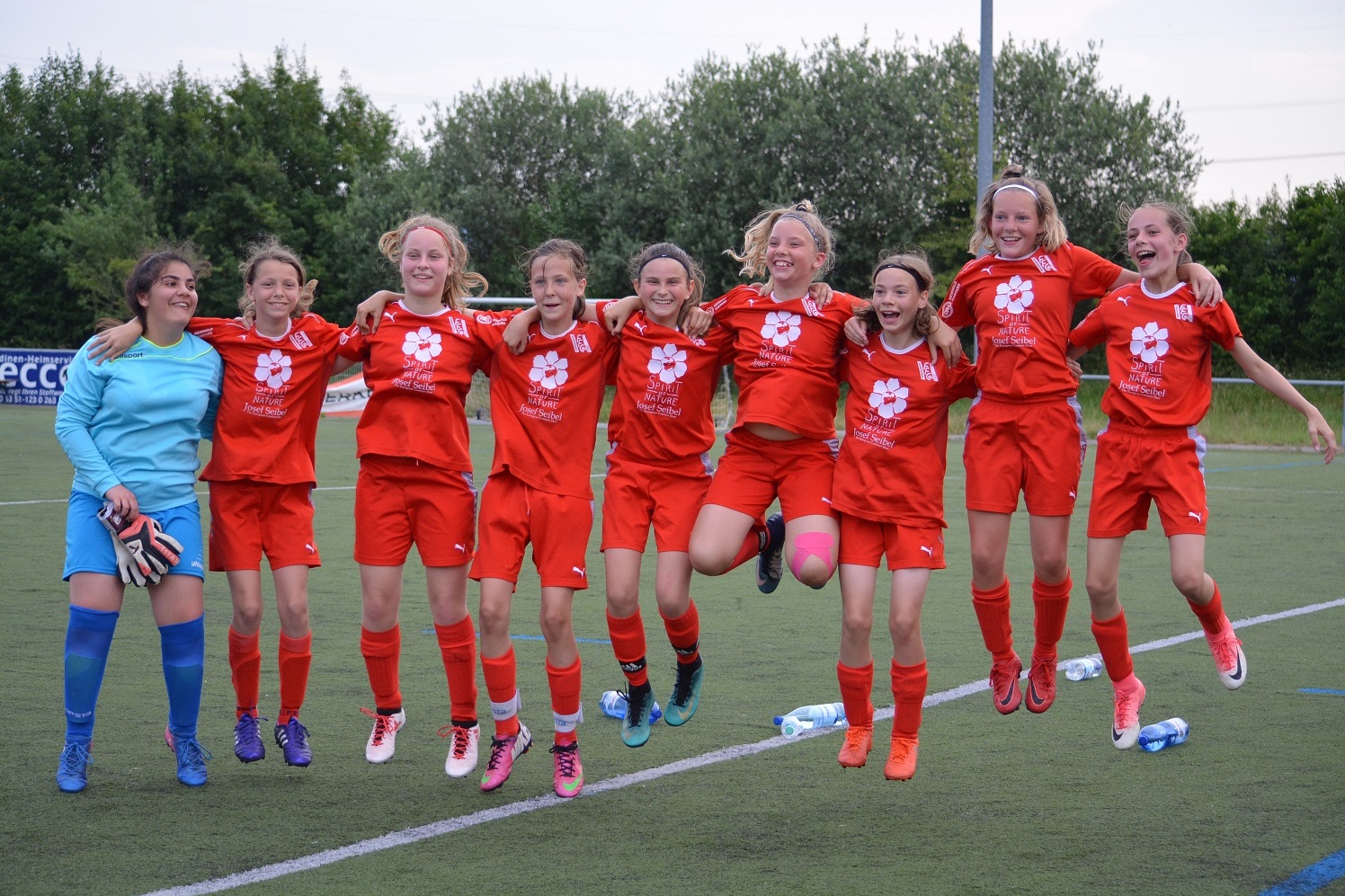Gruppe von neun M&auml;dchen in roten Trikots feiert gemeinsam auf einem Fu&szlig;ballfeld, einige springen vor Freude.