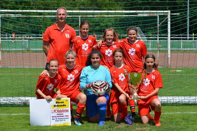 Sieben weibliche Fu&szlig;ballspielerinnen in roten Trikots stehen vor einem Tor, mit einem Pokal und einer Urkunde in der Mitte.