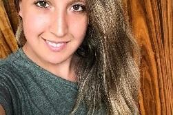 Portr&auml;t einer l&auml;chelnden Frau mit langen, gestuften Haaren vor einer Holzoberfl&auml;che. Sie tr&auml;gt ein graues T-Shirt.