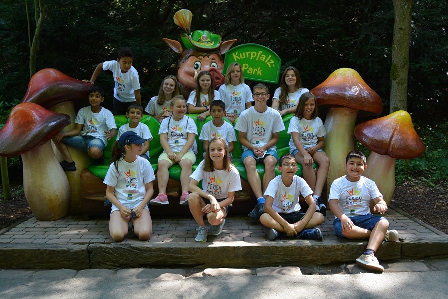 Gruppenzusammenstellung von Kindern in wei&szlig;en T-Shirts mit Aufschrift &bdquo;Ferien am Ch&ldquo; vor einer Pilzskulptur im Kurpfalz-Park.