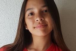 Portr&auml;t eines M&auml;dchens mit langen Haaren, das in einem roten Sporttrikot vor einer hellen Wand l&auml;chelt.