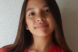 Portr&auml;t eines M&auml;dchens mit langen Haaren, das in einem roten Sporttrikot vor einer hellen Wand l&auml;chelt.