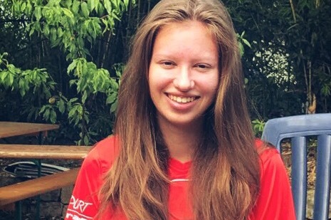 L&auml;chelnde junge Frau mit langen Haaren in rotem T-Shirt sitzt im Freien, umgeben von B&auml;umen und Holzst&uuml;hlen.