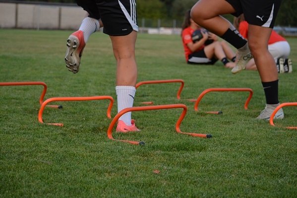 Sportlerinnen &uuml;berqueren orangefarbene H&uuml;rden auf einem gr&uuml;nen Rasenfeld w&auml;hrend eines Trainings.