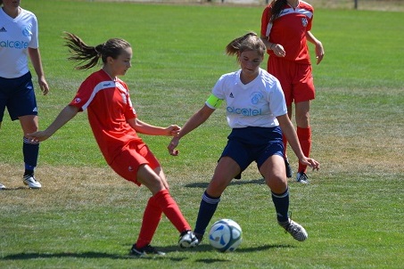 Zwei junge Fu&szlig;ballspielerinnen, eine in roter und eine in wei&szlig;-blauer Uniform, k&auml;mpfen um den Ball auf dem Spielfeld.