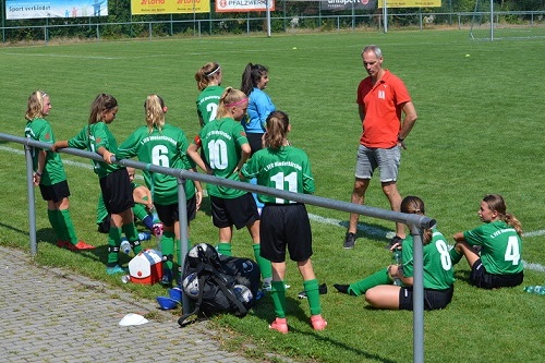 M&auml;dchenfu&szlig;ballmannschaft in gr&uuml;nen Trikots sitzt und steht auf dem Rasen, Trainer spricht mit ihnen.