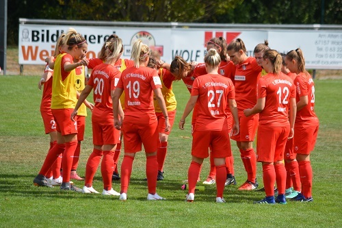 Frauenfu&szlig;ballteam in roten Trikots versammelt sich im Kreis auf dem Spielfeld f&uuml;r eine Besprechung.