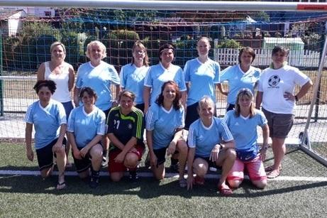 Gruppe von 14 Frauen in sportlichen Trikots steht vor einem Fußballtor auf einem Kunstrasenplatz.