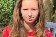 Portr&auml;t eines M&auml;dchens mit langen Haaren, das in einem roten Shirt vor gr&uuml;ner Vegetation sitzt.