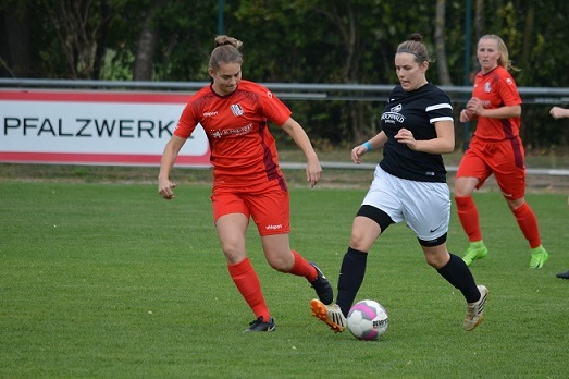 Zwei Fu&szlig;ballspielerinnen in roten und schwarzen Trikots k&auml;mpfen um den Ball auf einem Rasenplatz.