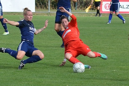 Zwei Fu&szlig;ballspielerinnen k&auml;mpfen um den Ball auf einem gr&uuml;nen Spielfeld; eine tr&auml;gt ein orangefarbenes Torwarttrikot.