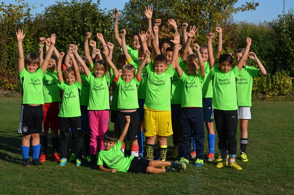 Gruppenshot von Kindern in leuchtend gr&uuml;nen T-Shirts, die mit erhobenen H&auml;nden auf einem Fu&szlig;ballplatz posieren.