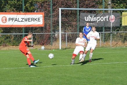 Spielszene mit einem Spieler in roter Kleidung, der auf zwei Abwehrspieler in wei&szlig;en Trikots mit einem Fu&szlig;ball zusprintet.