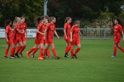 Eine Gruppe von acht Fu&szlig;ballspielerinnen in roten Trikots feiert einen Erfolg auf dem gr&uuml;nen Spielfeld.