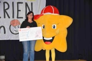 Frau h&auml;lt einen Scheck neben einem gelben Maskottchen mit rotem Hut auf einer B&uuml;hne. Hintergrund zeigt ein Banner.