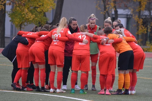 Eine Gruppe von Fu&szlig;ballspielerinnen in roten Trikots versammelt sich im Kreis auf einem Sportplatz, um sich zu besprechen.