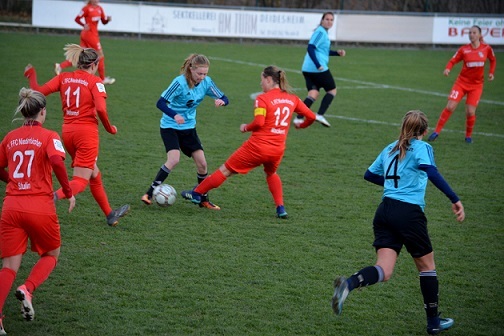 Zwei Gruppen von Fu&szlig;ballspielerinnen in roten und blauen Trikots k&auml;mpfen um den Ball auf einem Rasenplatz.