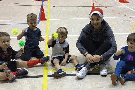 F&uuml;nf Kinder und eine Betreuerin sitzen auf dem Boden einer Sporthalle und halten bunte B&auml;lle in den H&auml;nden.