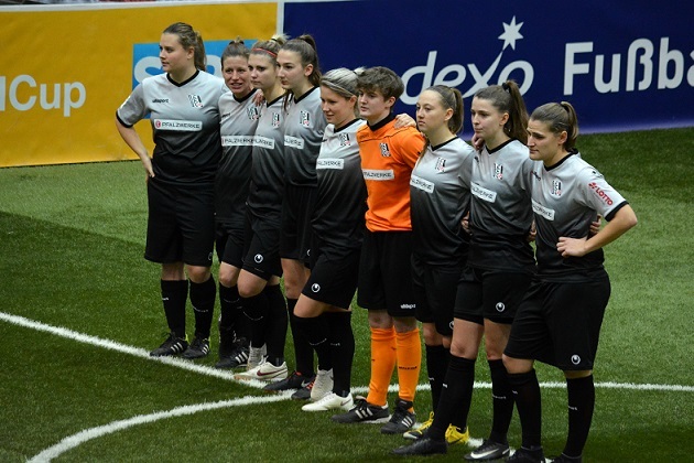 Mannschaft von Frauenfu&szlig;ballspielerinnen in schwarzen Trikots steht auf dem Feld vor einem Turnierbanner.