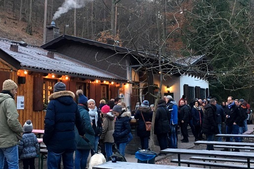 Menschen stehen an einer Holzschutzh&uuml;tte im Wald, w&auml;hrend Rauch aus dem Schornstein aufsteigt.