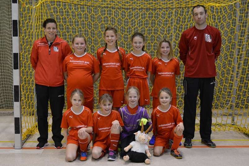 M&auml;dchen-Fu&szlig;ballmannschaft in roten Trikots steht vor einem Tor mit einem Trainer und einer Trainerin.