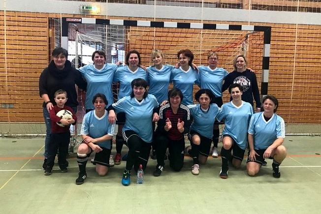 Gruppensnapshot einer Frauenfu&szlig;ballmannschaft in blauen Trikots mit einem Jungen und einer Fu&szlig;ball auf einem Hallenfeld.